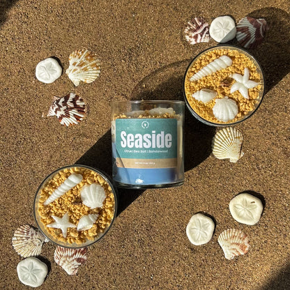 A candle named Seaside, with a beach-themed design, surrounded by seashells and sand on a sunny day.